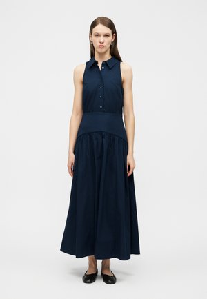 Ženska z dolgimi rjavimi lasmi, oblečena v temno modro obleko brez rokavov z gumbi in črnimi balerinkami, stoji pred belim ozadjem.