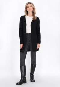 Zwarte lange cardigan van zachte brei stof, met open voorkant, lange mouwen, gecombineerd met een witte top, skinny grijze jeans en zwarte laarzen tot de knie.
