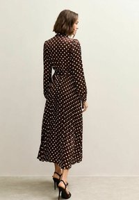 Bruine jurk met polka dots, lange mouwen en een ceintuur om de taille, met een vloeiende rok die tot halverwege de kuiten reikt, gemaakt van lichtgewicht stof.