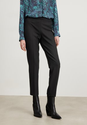 Mustat räätälöidyt housut, joissa on kapea istuvuus, yhdistetty sinivihreään kuvioituun puseroon. Ominaisuuksina sileä pinta ja nilkkaan ulottuvat mustat korolliset nilkkurit.