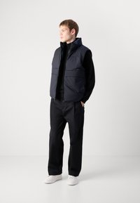 Giovane in dolcevita nera, gilet imbottito blu navy, pantaloni neri e sneakers bianche in piedi con le mani in tasca, che guarda a sinistra.