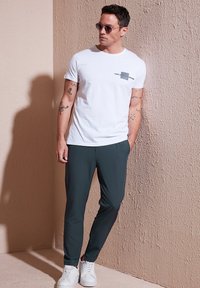Witte T-shirt met grafische print, donkergroene tapered broek en witte sneakers. Model staat tegen een gestructureerde beige muur.