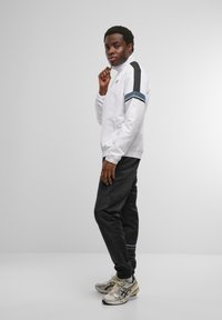 Vit zip-up jacka med svarta och teal-ränder, kombinerad med svarta byxor och silverfärgade sneakers. Slät tyg och åtsittande passform.