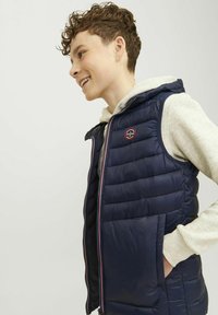 Temno modra puhasta jopica z quilted teksturo, poln zadrgo in kapuco. Ima logo obliž in stranski žep. Nosi se čez svetlo sivo kapuco.