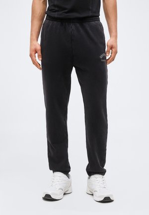 Man wearing black sweatpants and white sneakers, standing against a plain background with hands relaxed by sides.