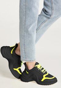 Zwarte sport sneakers met een gestructureerd oppervlak en zware zolen. Heldere gele accenten op de veters en zijpanelen. Draag met lichtblauwe jeans.