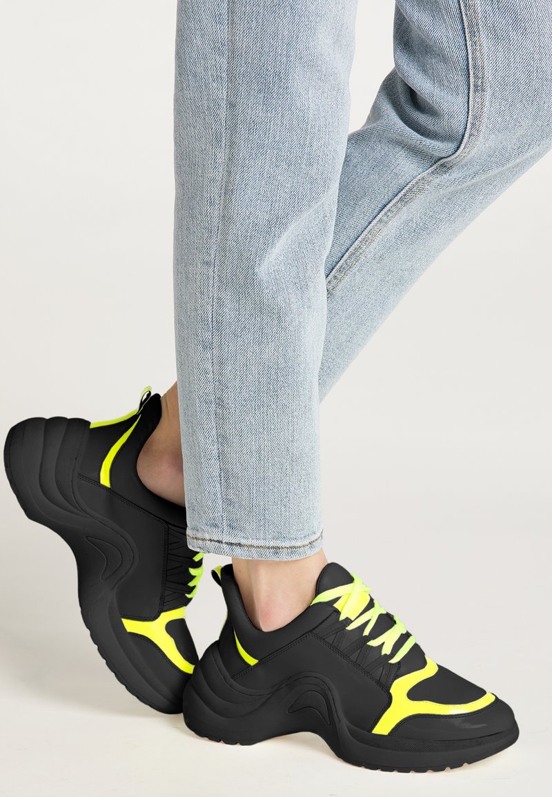 Zwarte sport sneakers met een gestructureerd oppervlak en zware zolen. Heldere gele accenten op de veters en zijpanelen. Draag met lichtblauwe jeans.