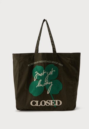Donkergroene draagtas met lange hengsels, met een grote groene klaver met vier blaadjes en tekst "Just get lucky," "European Heritage Since 1978," en "CLOSED."