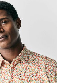 Camisa de manga corta con collar, con un fondo color crema y un patrón repetido de frutas amarillas y rojas, adornadas con hojas verdes.