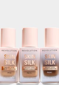 Tre frostat glasflaskor med foundation märkta "Revolution Skin Silk", med rosa lock och guldaccenter, med varierande nyansnummer.