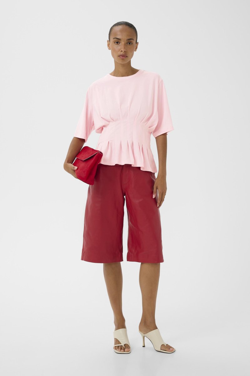 Kvinde iført en lyserød plisseret bluse, røde knælange shorts, hvide åbne sko med hæl, der holder en rød clutch mod en hvid baggrund.