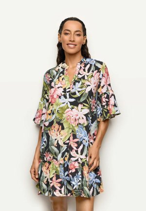 Femme souriante portant une robe noire fleurie avec des fleurs roses, bleues et vertes, debout devant un fond clair uni.