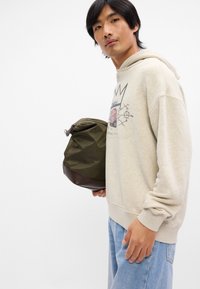 Πράσινη τσάντα υφάσματος με καφέ βάση, κλείσιμο με φερμουάρ και στρογγυλό σχήμα. Μοντέλο με μπεζ φούτερ και ανοιχτό μπλε τζιν, κρατώντας την τσάντα.