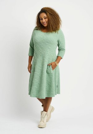 Grön texturerad klänning med vågig mönster, rund halsringning, trekvartsärmar, sidofickor; matchad med beige stövlar.