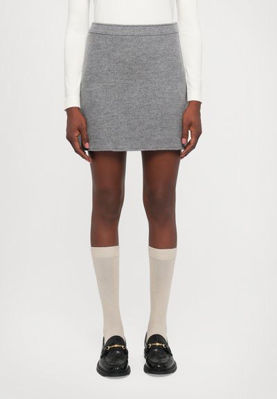 Model iført en grå uld mini-skjorte, hvid langærmet top, beige knæhøje sokker og sorte loafers med gyldne kæder.