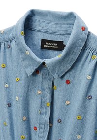 Chemise en denim bleu clair avec broderie florale en jaune, rouge, rose et violet. Présente un col pointu et une fermeture par boutons. Étiquette de marque visible.