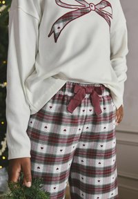 Vit ribbad långärmad tröja med en röd rosett-grafik, kombinerad med rutiga pyjamasbyxor med bordeaux, gröna och hjärtformade mönster.