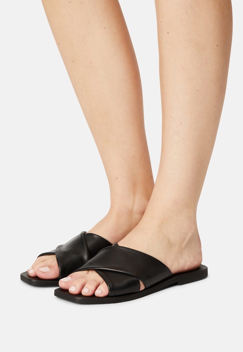 Pieds portant des sandales noires ouvertes à l'avant avec de larges brides en cuir croisées sur un fond clair.