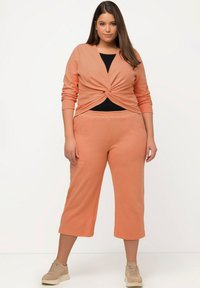 Top lavorato a maglia color pesco con dettaglio anteriore attorcigliato e maniche lunghe, abbinato a pantaloni cropped coordinati. Modella che indossa scarpe da ginnastica beige.