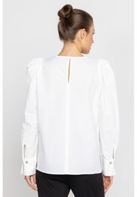 Biała bluzka z bufkami na rękawach, otwarciem na plecach w kształcie klucza i mankietami zapinanymi na guziki. Materiał wydaje się gładki i ma dopasowany krój.