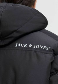 Czarna kurtka z kapturem, wykonana z teksturowanej tkaniny. Logo "JACK & JONES" jest wyraźnie widoczne w białym kolorze na plecach, na wysokości ramienia.