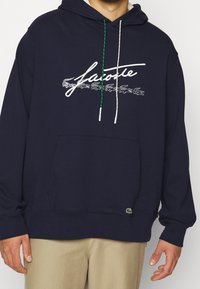 Tmavě modrá mikina s kapucí a klokaní kapsou, zdobená bílými a zelenými šňůrkami, vyšívaným logem a malým zeleným krokodýlem jako akcent.