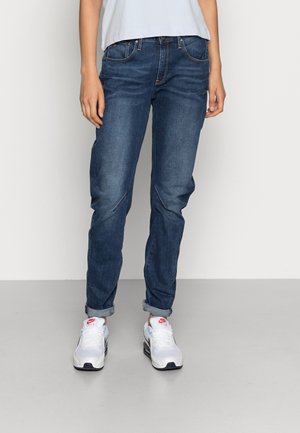 Denim jeans in mid-blue with a relaxed fit, featuring rolled cuffs, front pockets, and subtle stitching details. Paired with white sneakers.