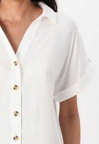 Camicia bianca a maniche corte con colletto a punte e bottoni frontali. Presenta una texture liscia e cuciture visibili sulle maniche.