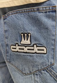 Hellblaue Jeans aus Denim mit einem schwarz-weißen gestickten Logo, das eine Krone und das Wort "dada" auf der Gesäßtasche zeigt.