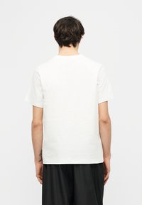 Άνδρας με σκουρόχρωμα μαλλιά που φαίνεται από την πίσω πλευρά, φοράει ένα απλό λευκό κοντομάνικο T-shirt και φαρδιά μαύρη παντελόνια, στέκεται μπροστά σε ανοιχτό γκρι φόντο.