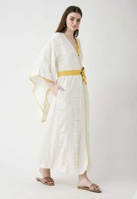 White textured maxi dress with geometric patterns, V-neck, yellow belt, and kimono sleeves, worn with brown slide sandals.