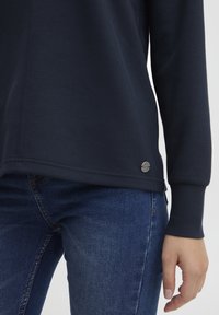 Mørkeblå langermet topp laget av mykt stoff, med rund skjæring og en sølvfarget logo på siden, satt sammen med blå denimjeans.