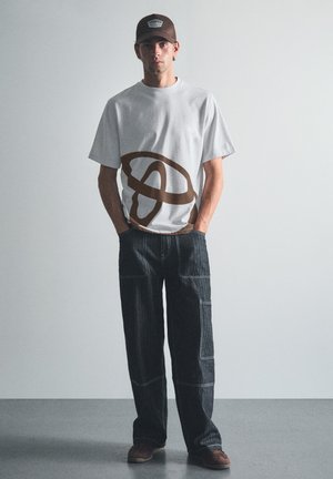 Jeune homme debout les mains dans les poches, portant un t-shirt blanc avec un motif abstrait marron, un jean foncé avec des détails de couture, une casquette marron et des chaussures.