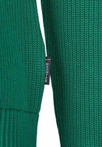 Grüner Strickpullover mit gerippten Bündchen und Säumen, mit einem sichtbaren Markenschild an der Seite. Die Textur ist dicht gewebt und glatt.