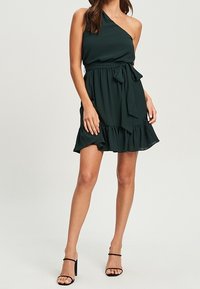 Ciemnozielona sukienka na jedno ramię z falbaniastym dołem, wysmukloną talią i pasującą kokardą. Wykonana z lekkiego, zwiewnego materiału.