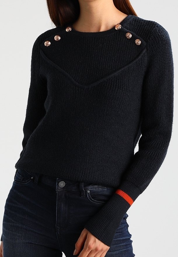Femme portant un pull en maille noir avec des boutons en cuivre sur les épaules et une bande orange au niveau du poignet de la manche droite.