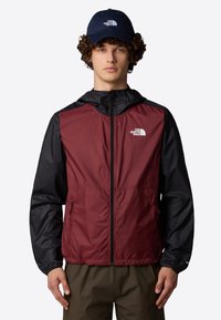 Giacca bordò e nera con zip frontale, cappuccio e tasche laterali. Presenta il logo sul lato sinistro del petto. I cappelli e i pantaloni sono in tonalità neutre.