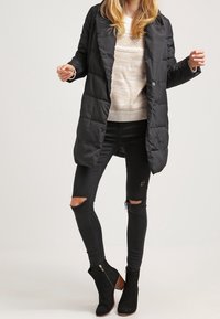 Musta toppatakki, jossa napit edessä, pitkät hihat, tikattu pinta, päällä vaalea neule ja revityt mustat farkut, yhdistetty nilkkureihin.