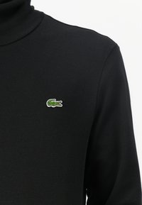 Pull col roulé noir en tissu doux, avec un petit logo de crocodile vert brodé sur la poitrine. Design simple, sans motifs supplémentaires.