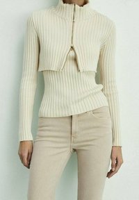 Suéter de canalé beige claro con cuello alto y cremallera frontal. Presenta mangas largas y un diseño ajustado, complementando pantalones beige.