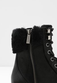 Sort ankelstøvle med faux fur krave, metallynlås og snørerdesign. Har en glat overflade og subtile stingdetaljer.