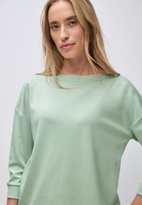 Lys grøn sweatshirt med bred halsudskæring, lange ærmer, ribbede manchetter og subtil tekstur på skuldrene. Glat stof og løs pasform.
