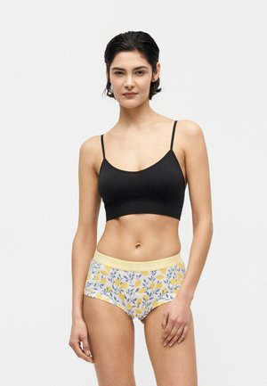 Moteris trumpais tamsiais plaukais, dėvinti juodą trumpą tanko viršūnę ir geltonius apatinius su gėlių raštu bei firminiu juosmeniu.