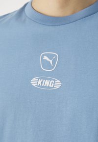 Šviesiai mėlynas medvilninis marškinėliai su apvaliomis iškirptėmis. Su baltu logotipu: Puma siluetas skydelyje ir "KING" horizontaliame elipse su juostelėmis.
