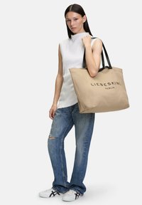 Beige Canvas-Shopper mit schwarzen Griffen, ausgestattet mit dem auffällig aufgedruckten Schriftzug "LIEBESKIND BERLIN". Das Modell trägt ein strukturiertes weißes Oberteil und verwaschene Jeans.