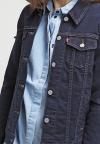 Granatowa dżinsowa kurtka z zapięciem na guziki z przodu, kieszeniami na piersi i usztywnionym kołnierzykiem, noszona na jasnoniebieskiej koszuli zapinanej na guziki.