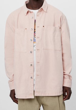 Person, die ein offenes, hellrosa Hemd über einem weißen T-Shirt mit Grafik und einer beigen Hose trägt, vor einem einfarbigen Hintergrund stehend.