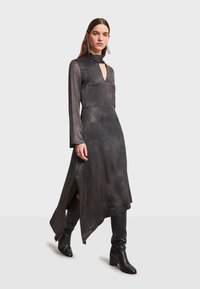 Dlhé čierne šaty s textúrovaným povrchom, hlbokým výstrihom do V, dlhými rukávmi, bočnými rozparkami a asymetrickým lemom. Skombinované s čiernymi čižmami po kolená.