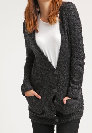 Femme portant un cardigan boutonné gris foncé texturé sur une chemise blanche, les mains dans les poches du cardigan.