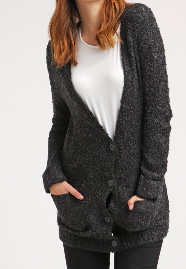Femme portant un cardigan boutonné gris foncé texturé sur une chemise blanche, les mains dans les poches du cardigan.
